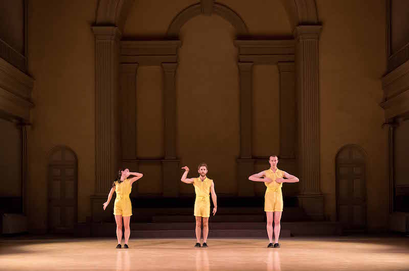 Three dancers in yellow rompers standing in a line. Their arms cover their face or eyes.